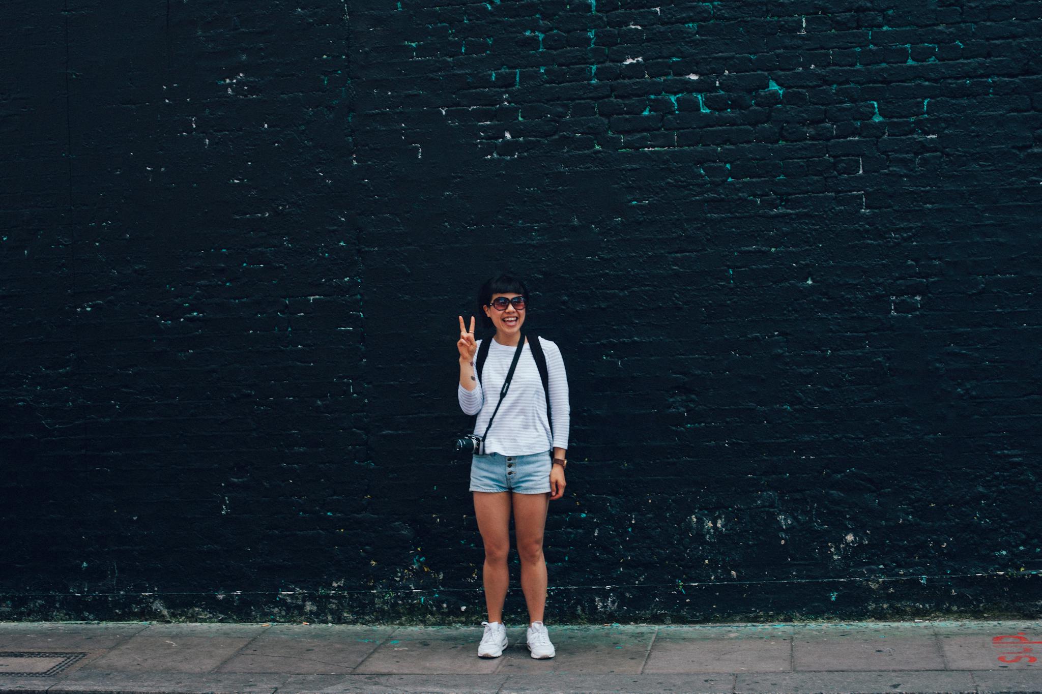 Smiling woman in sunglasses, posing with a peace sign against a brick wall.